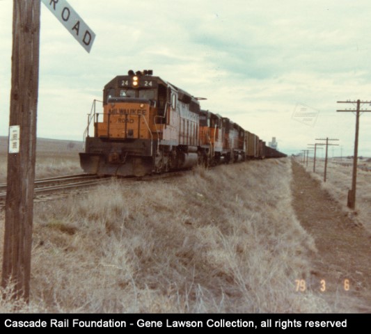 MILW 24 leading a westbound train at the west switch of the Ralston, WA siding (Bauman Road grade crossing) in 1979.  This may be a Marengo Turn, with a long string of empty hoppers returning to Utah through the UP interchange at Marengo.